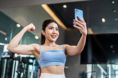 Mükemmel vücut giyimli genç Asyalı atletik kadın spor kıyafetleri giyerek spor salonunda spor yaparken başarılarını ve sonuçlarını göstermek için akıllı telefonuyla arkadaşlarına ya da ailesine video çağrısı yapıyor.