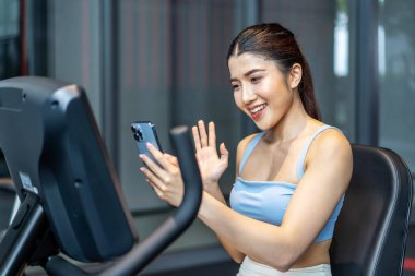 Mükemmel vücut giyimli genç Asyalı atletik kadın spor kıyafetleri giyerek spor salonunda spor yaparken başarılarını ve sonuçlarını göstermek için akıllı telefonuyla arkadaşlarına ya da ailesine video çağrısı yapıyor.