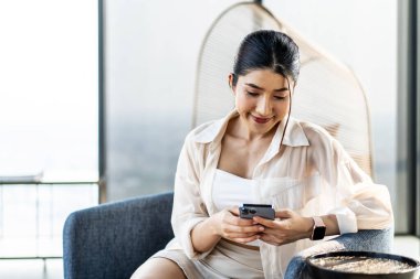 Gülümseyen Asyalı kadın evde otururken akıllı telefon kullanıyor. Sosyal medyayı izlemek. Portre, eğlence, iletişim, çalışma veya öğrenim, bağlantı, mobil uygulamalar, yaşam tarzı kavramı.