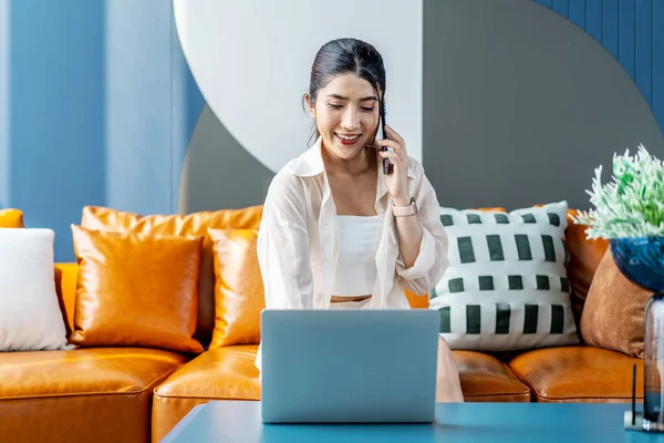 Happy young asian woman freelancer sit on comfy couch work at laptop talk on phone to friend customer distracted from online job, contact and consulting with customer in living room for ecommerce