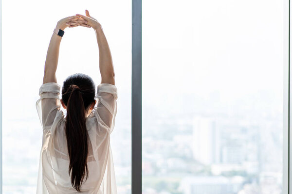 Young asian woman standing in front of big window, stretching arms and back, enjoying good morning time in office. Happy attractive millennial girl feeling no stress and peace of mind concept