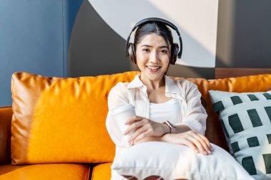 Haftasonu rahatla, kulaklıklı genç Asyalı kadın koltukta oturup en sevdiği müziği dinliyor ve kanepede kahve içiyor, rahatlama ve meditasyon kavramının tadını çıkarıyor.