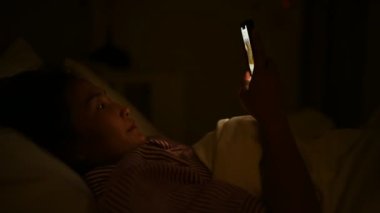 Asyalı kadın gece yatakta akıllı telefonuyla oyun oynuyor. Taylandlılar. Sosyal medyaya bağımlılık