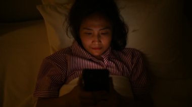 Asyalı kadın gece yatakta akıllı telefonuyla oyun oynuyor. Taylandlılar. Sosyal medyaya bağımlılık