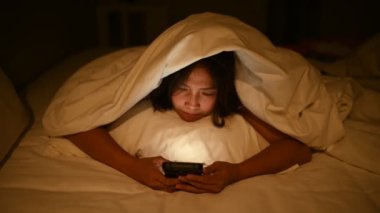 Asyalı kadın gece yatakta akıllı telefonuyla oyun oynuyor. Taylandlılar. Sosyal medyaya bağımlılık