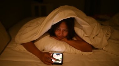 Asyalı kadın gece yatakta akıllı telefonuyla oyun oynuyor. Taylandlılar. Sosyal medyaya bağımlılık