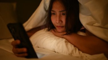 Asyalı kadın gece yatakta akıllı telefonuyla oyun oynuyor. Taylandlılar. Sosyal medyaya bağımlılık