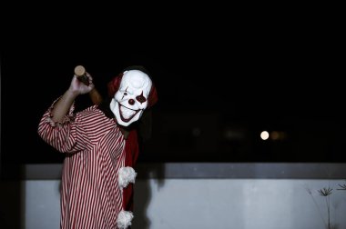 Asyalı yakışıklı adam gece olay yerinde silahlı palyaço maskesi giyiyor, Cadılar Bayramı festivali konsepti, turuncu kıyafetli bir katilin korkunç fotoğrafı, şeytani palyaço avcısı.