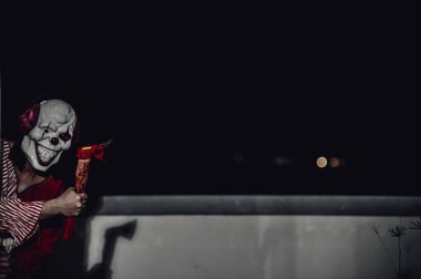 Asyalı yakışıklı adam gece olay yerinde silahlı palyaço maskesi giyiyor, Cadılar Bayramı festivali konsepti, turuncu kıyafetli bir katilin korkunç fotoğrafı, şeytani palyaço avcısı.