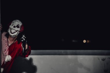 Asyalı yakışıklı adam gece olay yerinde silahlı palyaço maskesi giyiyor, Cadılar Bayramı festivali konsepti, turuncu kıyafetli bir katilin korkunç fotoğrafı, şeytani palyaço avcısı.