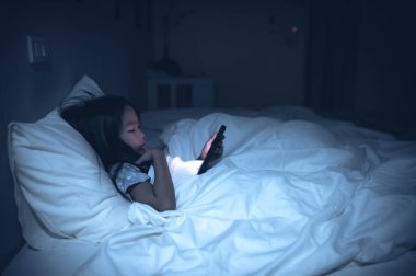 Asyalı çocuk gece yatakta akıllı telefondan oyun oynuyor. Sosyal medya bağımlısı kız.