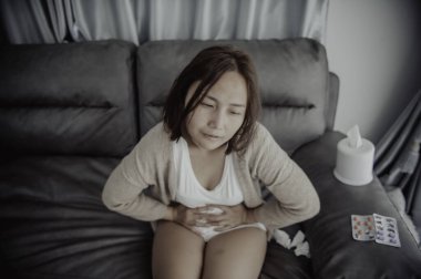 Asyalı hasta kadın kanepede oturur, evde kalır, kadın kendini kötü hisseder, uzanır ve dinlenmek ister, karın ağrısı, regl sancısı...