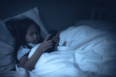 Asyalı çocuk gece yatakta akıllı telefondan oyun oynuyor. Sosyal medya bağımlısı kız.