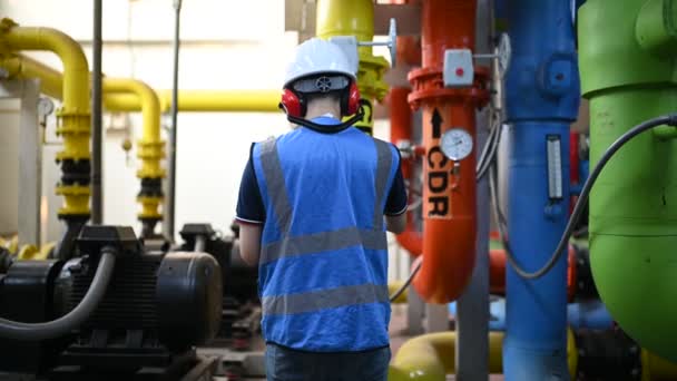 暖房工場のメンテナンス技術者 石油化学労働者は工場内のガスパイプラインと石油パイプラインの運営を監督している エンジニアは補聴器を多くのパイプを備えた部屋に置きます — ストック動画