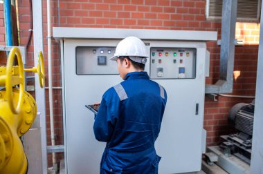 Büyük bir fabrikada çalışan genç bir mühendis koruyucu üniformalı bir teknisyen ve borulardaki ısıyı kontrol eden bir baret.