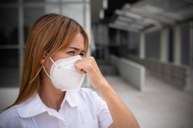 Asyalı kadın N95 maskesi takıyor 2.5 numaralı toz bulutunu korumak için, ekoloji konsepti. Alerji, baş ağrısı. Şehirde hava tehlikesi.