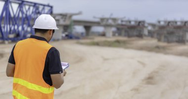 Asyalı mühendislerden oluşan bir grup inşaat alanındaki çalışmalar hakkında konuşuyor, müteahhit ekip binanın tasarımını tartışıyor, üç işçi dışarıda çalışıyor..