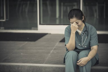 Depresyonda olan Asyalı kadın hemşire yüz maskesi giyiyor mavi üniforma hastane zemininde oturuyor, genç kadın doktor sıkı çalışmaktan strese giriyor.
