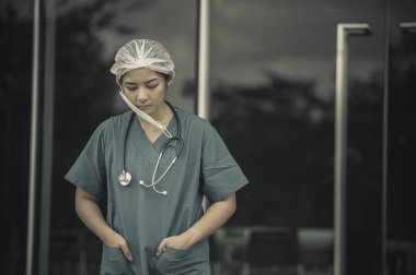 Depresyonda olan Asyalı kadın hemşire yüz maskesi giyiyor mavi üniforma hastane zemininde oturuyor, genç kadın doktor sıkı çalışmaktan strese giriyor.
