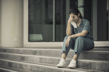Depresyonda olan Asyalı kadın hemşire yüz maskesi giyiyor mavi üniforma hastane zemininde oturuyor, genç kadın doktor sıkı çalışmaktan strese giriyor.