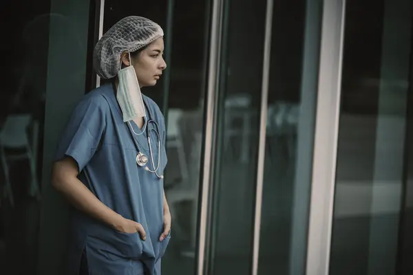 Tired depressed female asian scrub nurse wears face mask blue uniform sits on hospital floor. Young woman doctor stressed from hard work