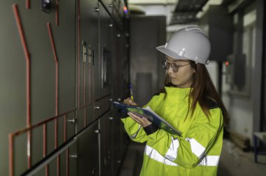 Asyalı elektrik mühendisi kontrol odasındaki elektrik sistemini kontrol etmek ve izlemek için pano tutuyor. Tayland teknisyeni ve çalışanlar