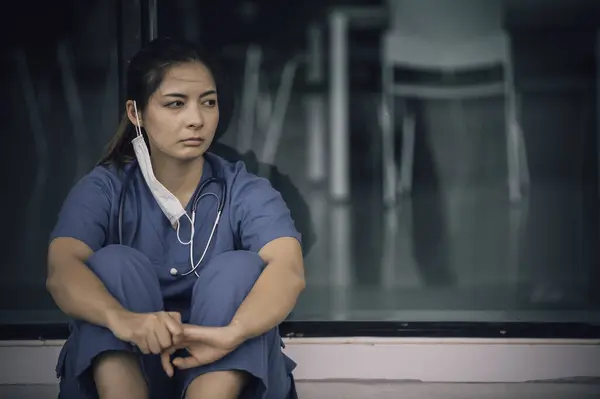 Tired depressed female asian scrub nurse wears face mask blue uniform sits on hospital floor. Young woman doctor stressed from hard work