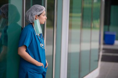 Depresyonda olan Asyalı kadın hemşire yüz maskesi giyiyor mavi üniforma hastane zemininde oturuyor, genç kadın doktor sıkı çalışmaktan strese giriyor.