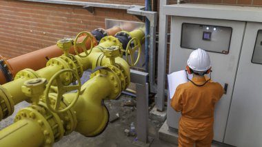 Büyük bir fabrikada çalışan genç bir mühendis koruyucu üniformalı bir teknisyen ve borulardaki ısıyı kontrol eden bir baret.