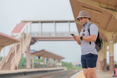 Sırt çantalı yakışıklı genç adam, tren peronunda, turizmde ve yazın seyahat ederken telefonu kullanıyor. Öğrenciler için tatiller.