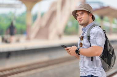 Sırt çantalı yakışıklı genç adam, tren peronunda, turizmde ve yazın seyahat ederken telefonu kullanıyor. Öğrenciler için tatiller.