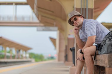 Sırt çantalı yakışıklı genç adam, tren peronunda, turizmde ve yazın seyahat ederken telefonu kullanıyor. Öğrenciler için tatiller.