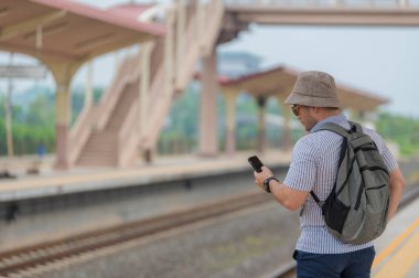 Sırt çantalı yakışıklı genç adam, tren peronunda, turizmde ve yazın seyahat ederken telefonu kullanıyor. Öğrenciler için tatiller.