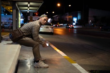 An Asian Man Waiting for a Bus After Working Late, Using His Smartphone at a Busy City Bus Stop, Capturing the Struggles of Urban Life, Overwork, and the Nightly Commute in a Chaotic Capital clipart