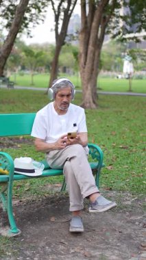 Kulaklıklı son sınıf öğrencisi bankta oturuyor ve parkta akıllı telefon kullanıyor.
