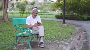 Kulaklıklı son sınıf öğrencisi bankta oturuyor ve parkta akıllı telefon kullanıyor.