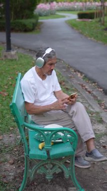 Kulaklıklı son sınıf öğrencisi bankta oturuyor ve parkta akıllı telefon kullanıyor.
