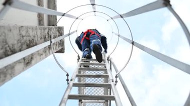 Miğferli ve koruyucu kıyafetli bir işçi metal merdivene tırmanıyor.