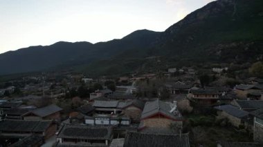 Gün batımında Baisha Antik Kasabası 'nın üzerinde süzülen video. Çatıları vadi boyunca uzanan geleneksel evler. Dağlar ufku kucaklıyor 