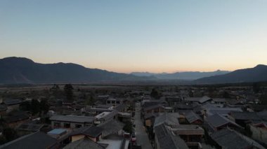 Gün batımında Baisha Antik Kasabası 'nın üzerinde süzülen video. Çatıları vadi boyunca uzanan geleneksel evler. Dağlar ufku kucaklıyor 