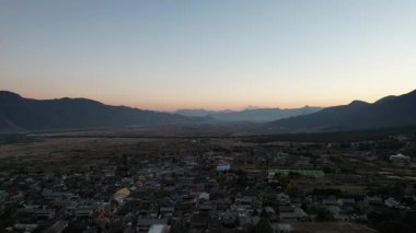 Gün batımında Baisha Antik Kasabası 'nın üzerinde süzülen video. Çatıları vadi boyunca uzanan geleneksel evler. Dağlar ufku kucaklıyor 