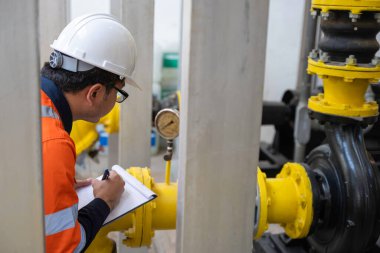 Kazan dairesinde çalışan gözlüklü Asyalı mühendis ısıtma sistemi ekipmanlarının teknik verilerini kontrol ediyor.