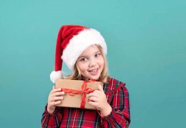 Noel Baba şapkalı çocuk mavi arka planda hediye kutusunu tutuyor. Noel kostümü giymiş, elinde hediyeyle beyaz çocuk portresi. Mutlu küçük bebek yeni yıl konsepti.