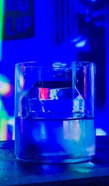 Geometrik şekilli buzlu cam bardak, mavi ve yeşil ışığın arka planında, bir bar tezgahında buzun yeşil ve kırmızı yansımaları vardır..