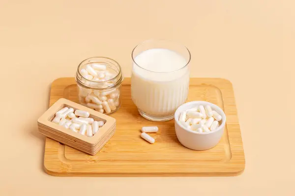 stock image Lactase enzyme tablets in a wooden box and in a ceramic white plate, in a glass jar, a glass of milk on wooden desk on beige background. Lactose intolerance. Dairy products containing lactose