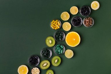 Yeşil arkaplandaki doğal vitaminlerin kaynağı olarak birçok beslenme takviyesi, vitamin ve mineral, renkli haplar ve küçük kaselerde bulunan tabletler, kivi, ıhlamur ve portakaldır. Vitaminler ve meyveler. 