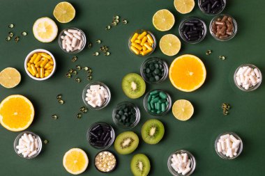 Yeşil arkaplandaki doğal vitaminlerin kaynağı olarak birçok beslenme takviyesi, vitamin ve mineral, renkli haplar ve küçük kaselerde bulunan tabletler, kivi, ıhlamur ve portakaldır. Vitaminler ve meyveler. 