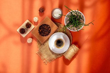 Ahşap masada bir fincan sıcak lezzetli kahve kahve, kahve çekirdekleri, mum ve ev bitkisi yukarıdan güzel bir kompozisyonda güneş ışığında turuncu arka planda. Amerikalı, espresso ve kavrulmuş kahve taneleri.