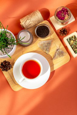 Ahşap masada bir kupa sıcak çay, cam kaselerde bitki çayı yaprakları, mum ve ev bitkisi yukarıdan güzel bir kompozisyonda, turuncu arka planda güneş ışığı altında..