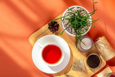 Ahşap masada bir kupa sıcak çay, cam kaselerde bitki çayı yaprakları, mum ve ev bitkisi yukarıdan güzel bir kompozisyonda, turuncu arka planda güneş ışığı altında..